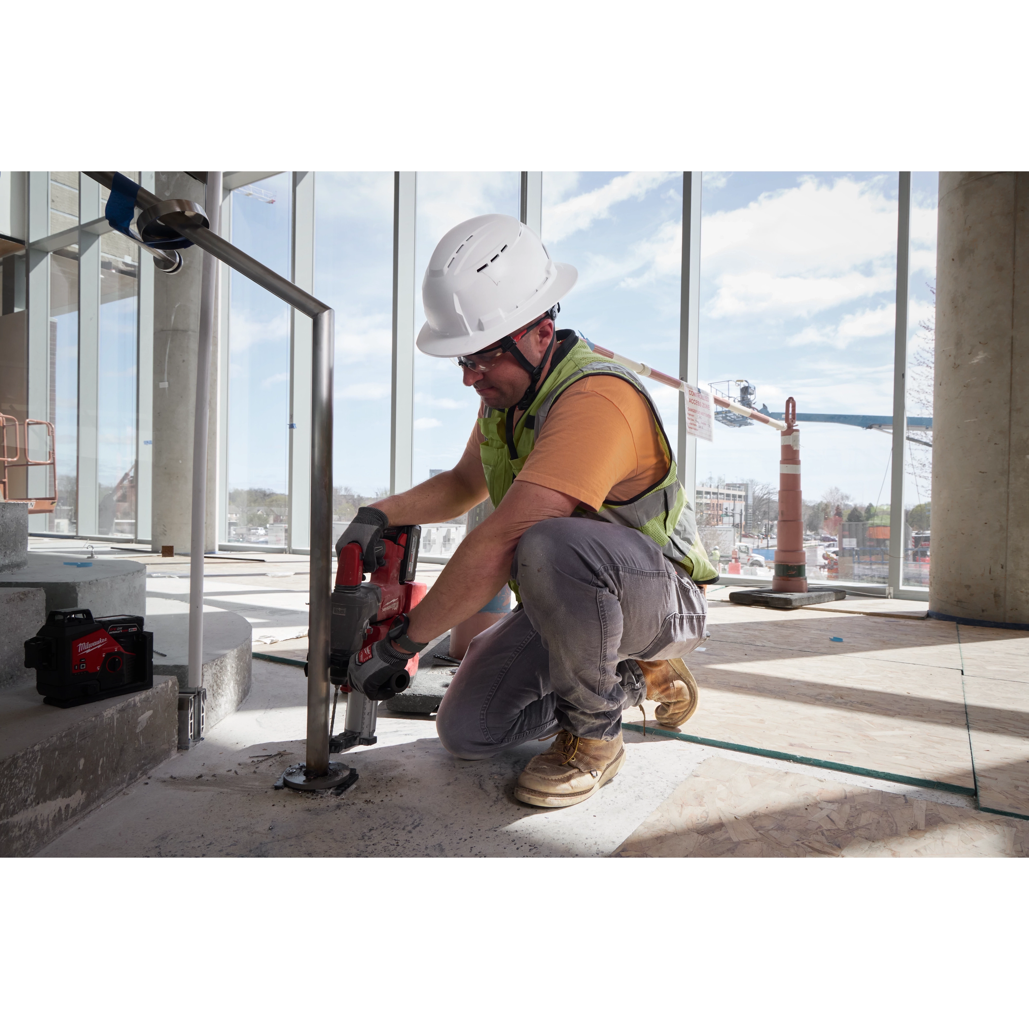A construction worker wears a BOLT™ White Full Brim Vented Safety Helmet – Type 2, Class C and yellow vest while operating a power tool inside a partially finished building. The background shows large windows with an outdoor view of construction equipment and safety cones.