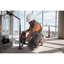 A construction worker wears a BOLT™ White Full Brim Vented Safety Helmet – Type 2, Class C and yellow vest while operating a power tool inside a partially finished building. The background shows large windows with an outdoor view of construction equipment and safety cones.