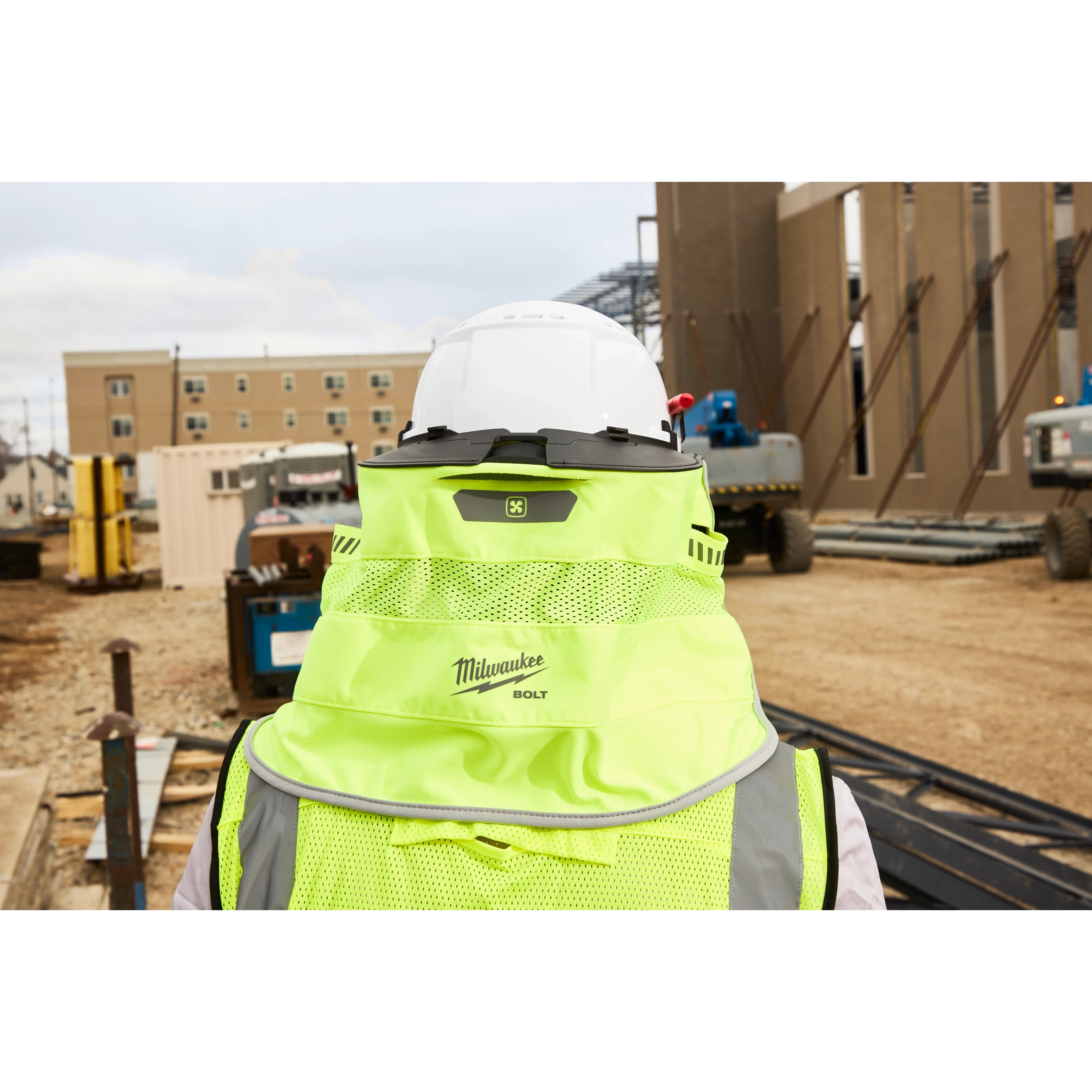 A construction worker wearing a neon yellow Milwaukee safety vest and a white hard hat is viewed from the back. The construction site features partially built structures, equipment, and building materials scattered around. Multi-story buildings are visible in the background.