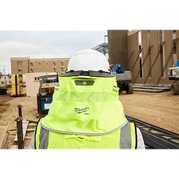 A construction worker wearing a neon yellow Milwaukee safety vest and a white hard hat is viewed from the back. The construction site features partially built structures, equipment, and building materials scattered around. Multi-story buildings are visible in the background.