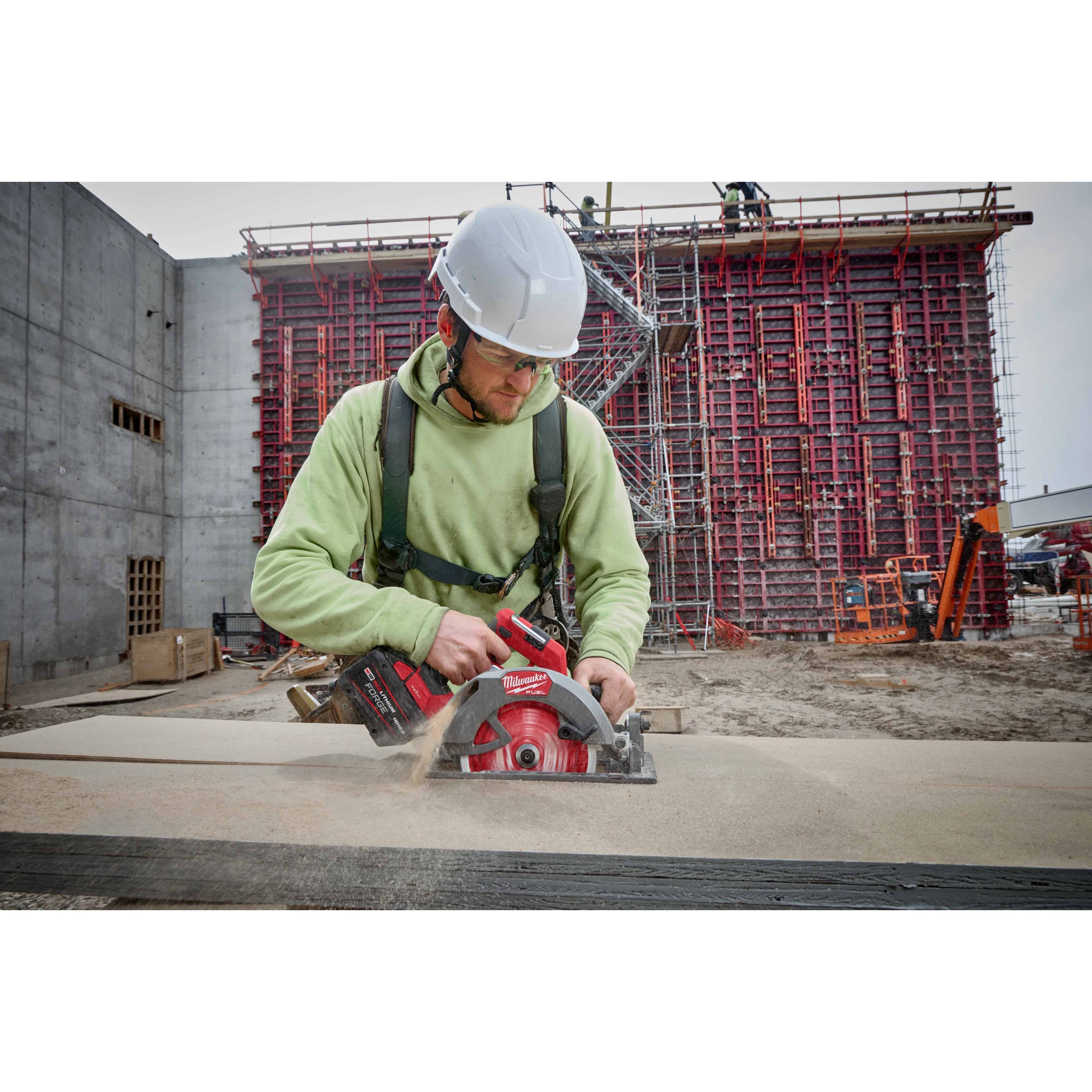 A construction worker uses the M18™ FUEL™ 7-1/4" Circular Saw w/ONEKEY™ to cut wood on a job site. The worker wears a green safety harness and operates the red and black saw that creates sawdust in the air. A large scaffold structure is built in the background.