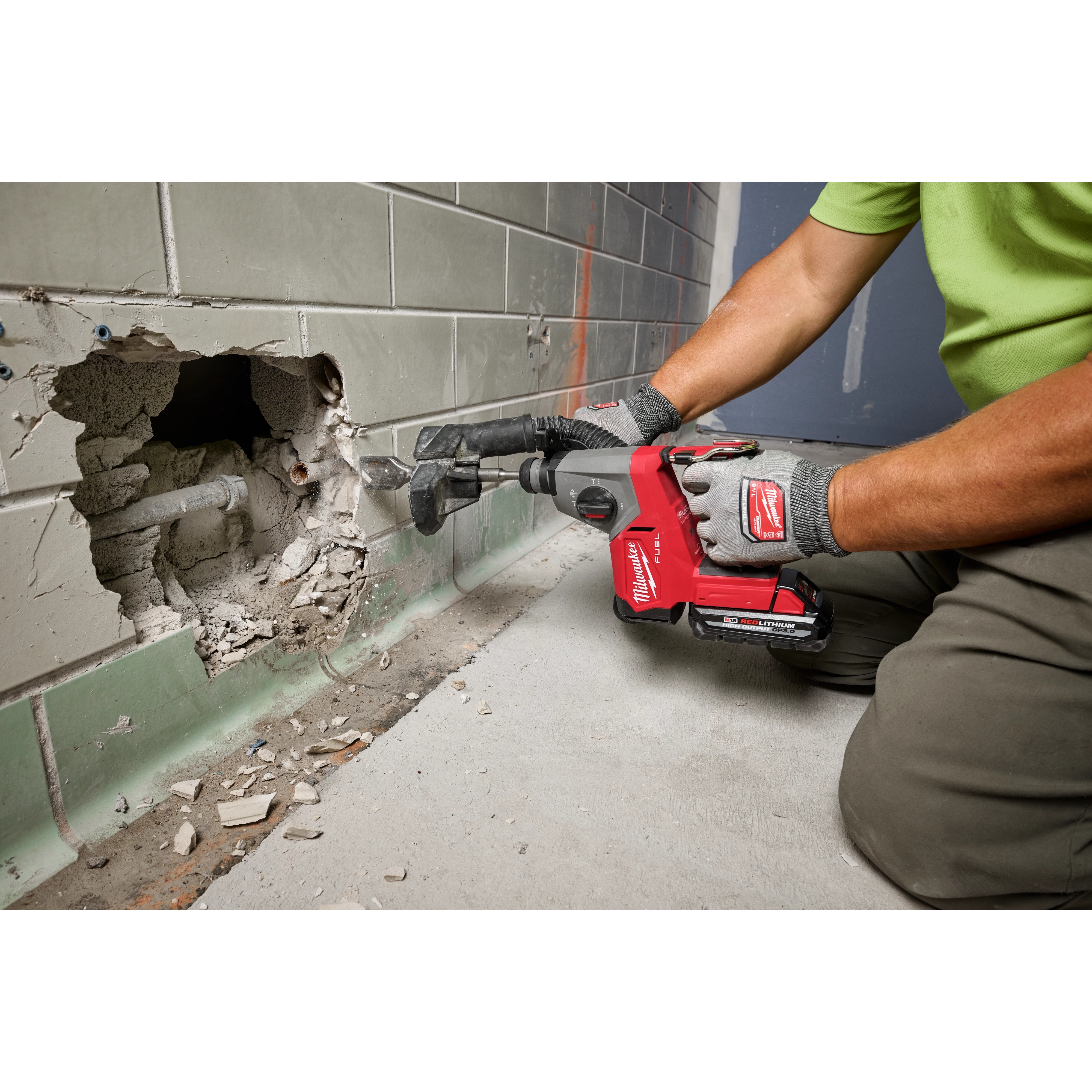 A person in work gloves uses the M18 FUEL™ 5/8" SDS Plus Rotary Hammer with Dust Extractor Kit to create a hole in a tiled wall, revealing pipes. The tool features a HAMMERVAC™ 5/8" Dedicated Dust Extractor for dust collection.