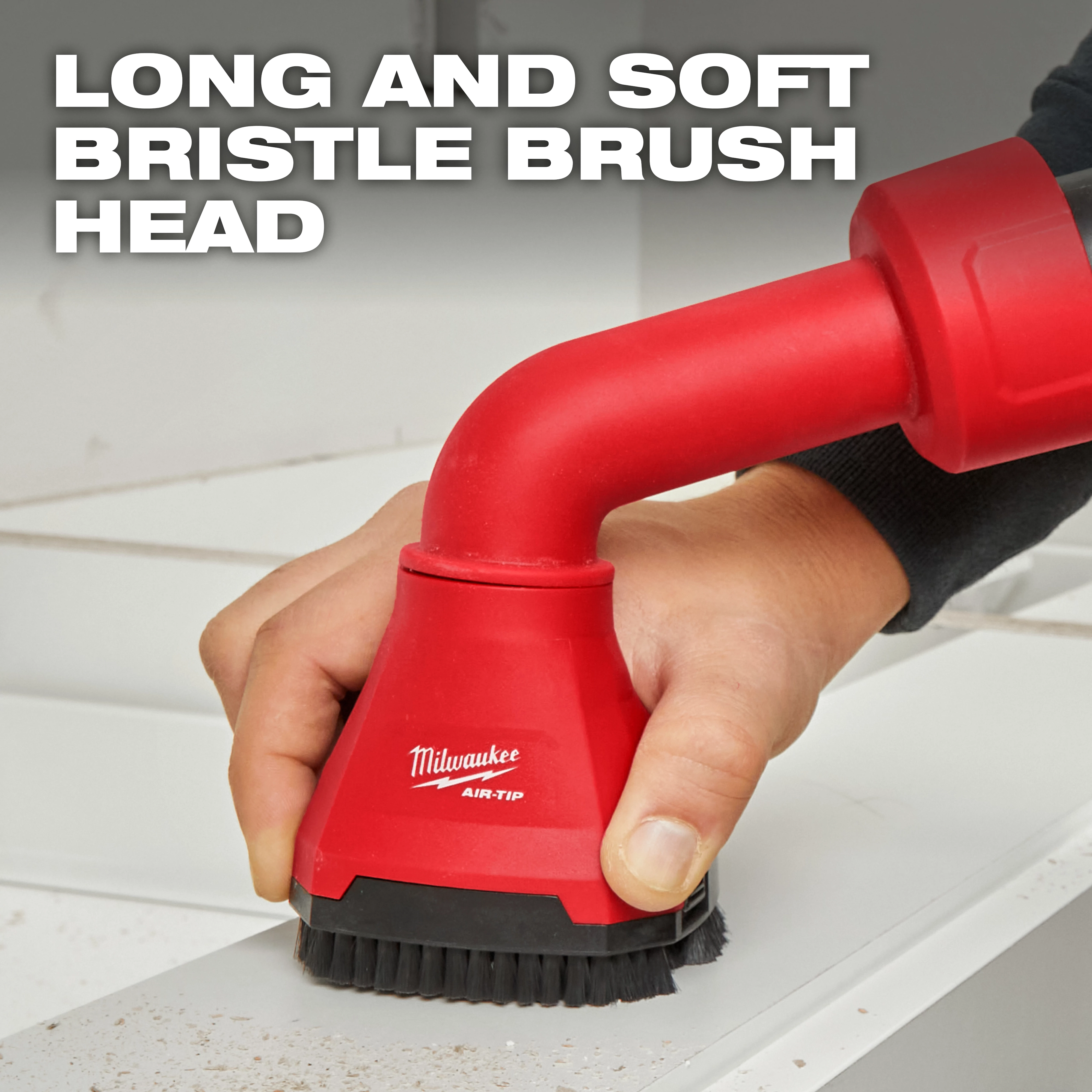 A hand using a Milwaukee AIR-TIP™ Swiveling Palm Brush with a long and soft bristle brush head to clean a dusty surface. The red brush attachment is connected to a vacuum hose. Text on the image reads, "LONG AND SOFT BRISTLE BRUSH HEAD."