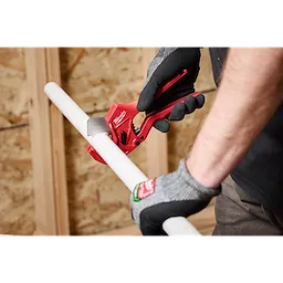 Person using 1-5/8" Ratcheting Pipe Cutter to cut a white PVC pipe, wearing black and gray gloves, with a wood background.
