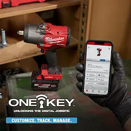 Gloved hand holds a phone showing M18 FUEL 3/4" High Torque Impact Wrench w/ ONE-KEY Kit beside the wrench on a workbench.