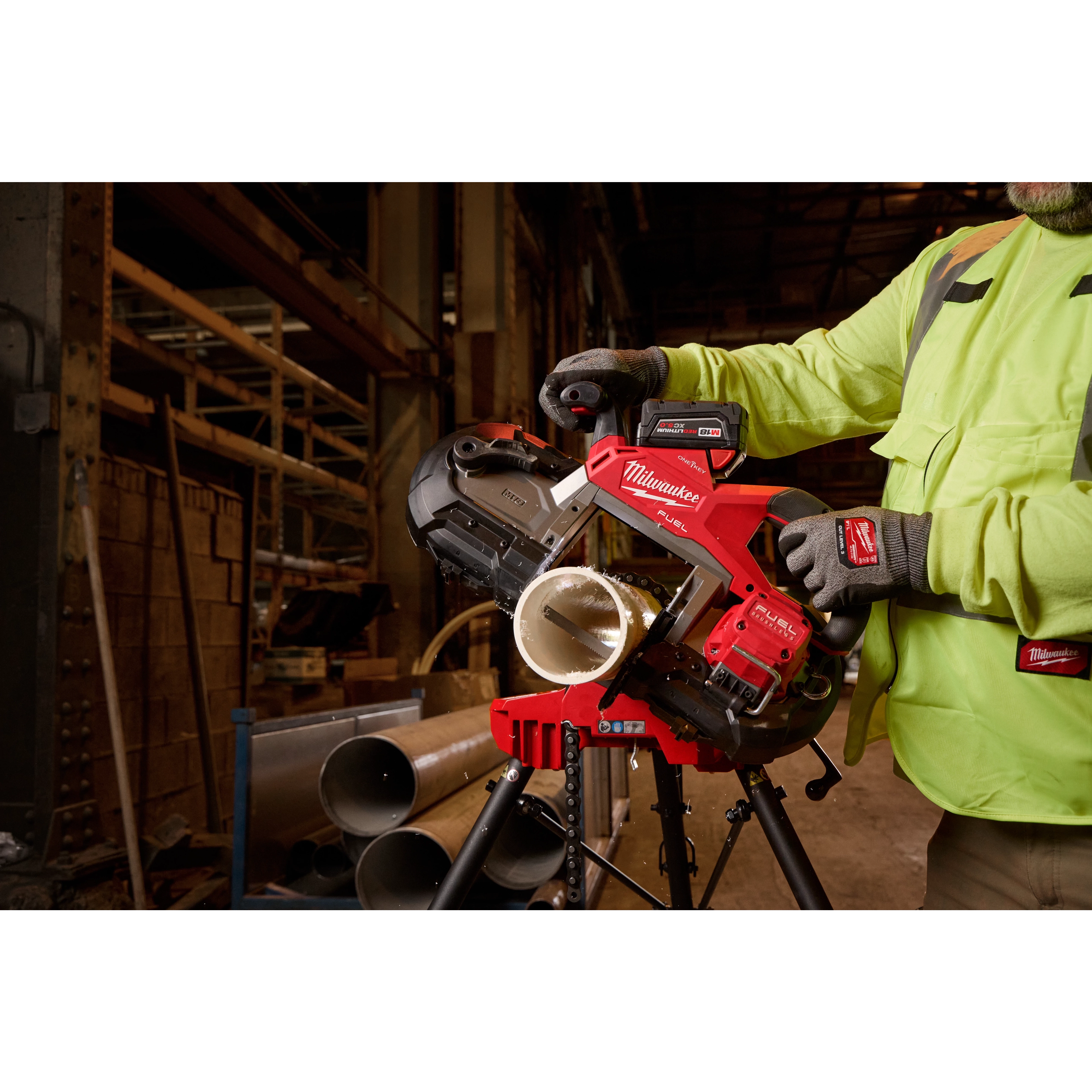 A worker operating the M18 FUEL™ Deep Cut Dual Trigger Band Saw w/ ONE-KEY™ to cut a metal pipe. The saw is red and black, and it is positioned on a workbench in an industrial setting. The worker is wearing safety gear, including gloves and a high-visibility jacket.