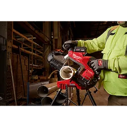 A worker operating the M18 FUEL™ Deep Cut Dual Trigger Band Saw w/ ONE-KEY™ to cut a metal pipe. The saw is red and black, and it is positioned on a workbench in an industrial setting. The worker is wearing safety gear, including gloves and a high-visibility jacket.