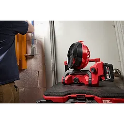 M18 Brushless Scissor Lift Fan in a workshop, placed on a red and black surface, with a person working in the background.