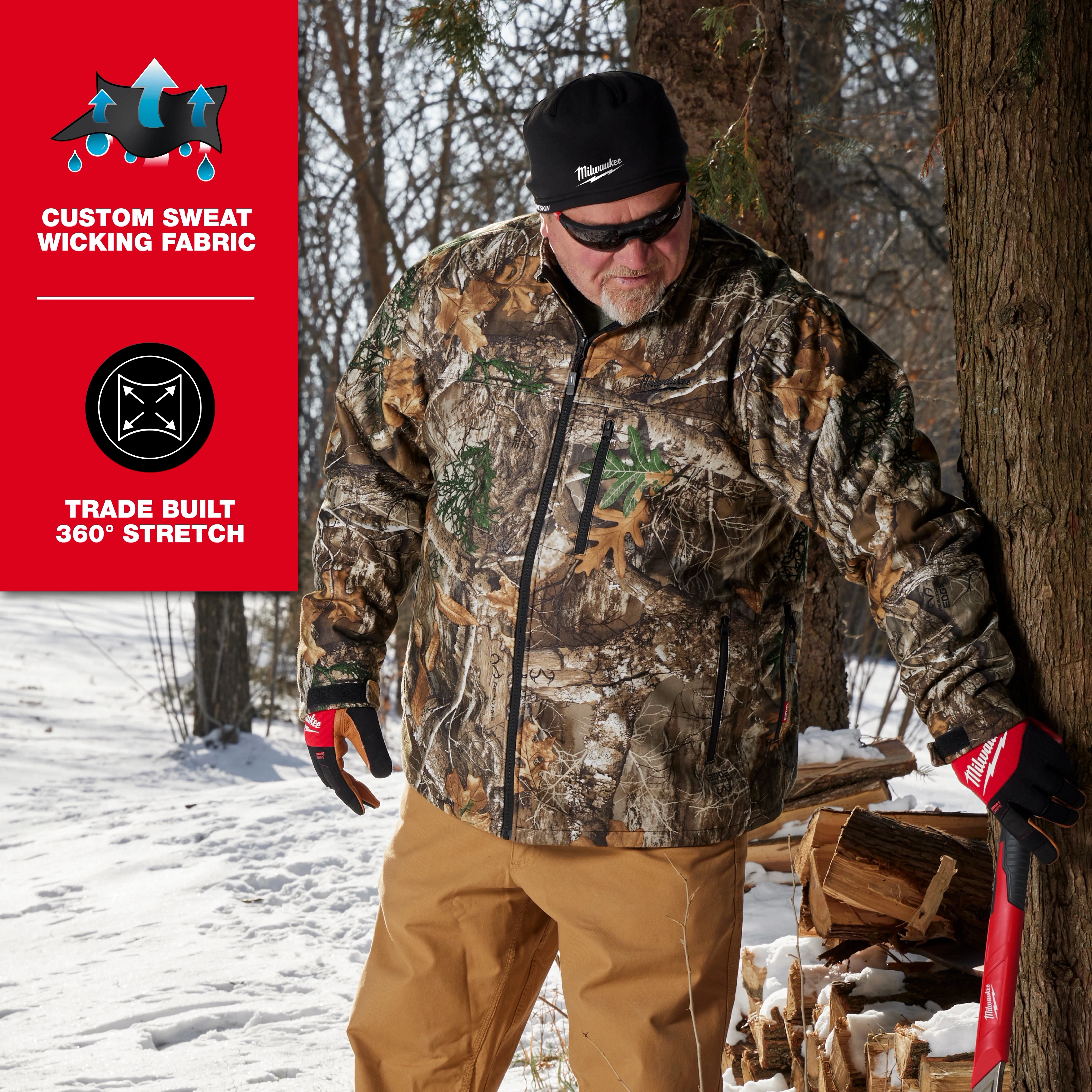 A person wearing camo jacket, tan pants, and red gloves stands beside a tree in snowy woods. The black WORKSKIN™ Low-Profile Beanie is on their head. Text on the image highlights "Custom Sweat Wicking Fabric" and "Trade Built 360° Stretch."