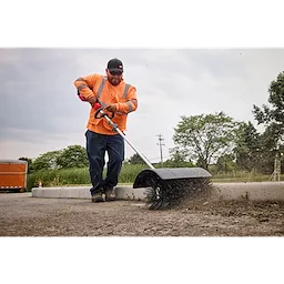 Worker in an orange safety vest using a street sweeper on a road, surrounded by greenery and traffic cones.