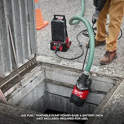 A worker operates the MX FUEL™ 1HP 2" Submersible Pump, lowering it into a water-filled pit at a construction site. The equipment is connected to a portable pump power base. Text at the bottom reads "MX FUEL™ portable pump power base & battery pack (not included) required for use."