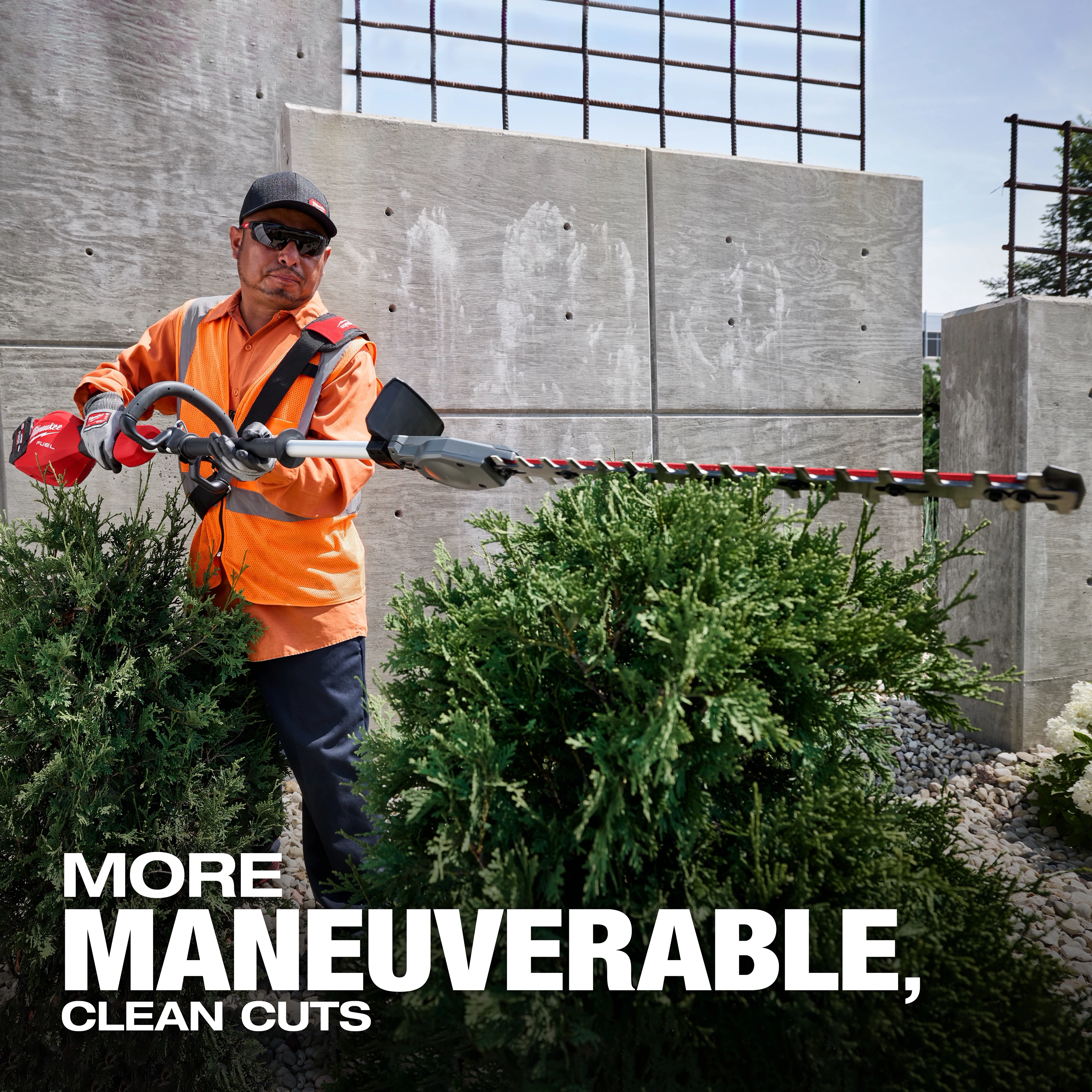 A person wearing an orange safety vest uses a hedge trimmer to cut green shrubs