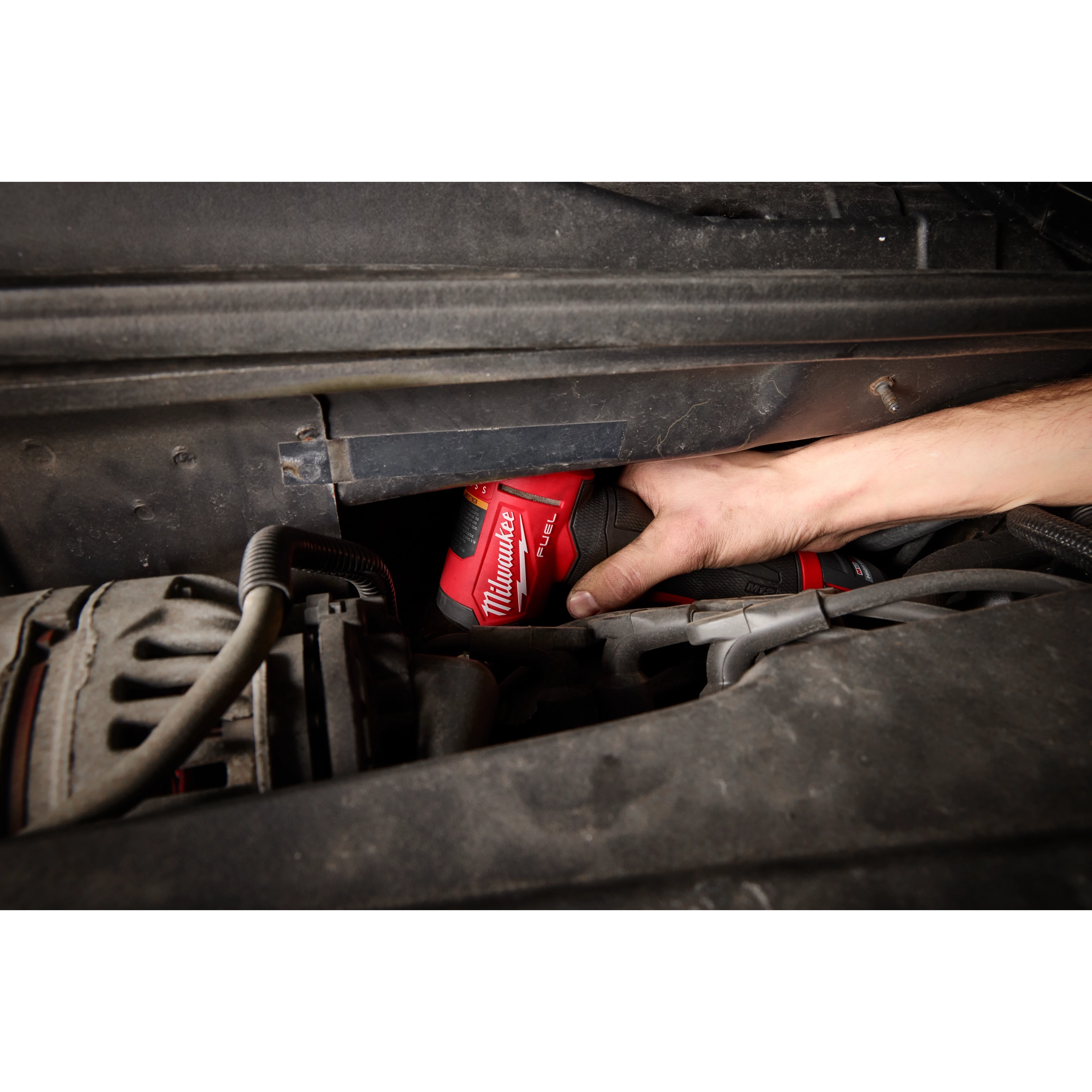 A person uses the M12 FUEL™ 1/4" Right Angle Die Grinder to work in a tight space under a vehicle's hood. The red tool is visible, and the hand holding it is reaching into the engine area.