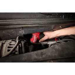 A person uses the M12 FUEL™ 1/4" Right Angle Die Grinder to work in a tight space under a vehicle's hood. The red tool is visible, and the hand holding it is reaching into the engine area.