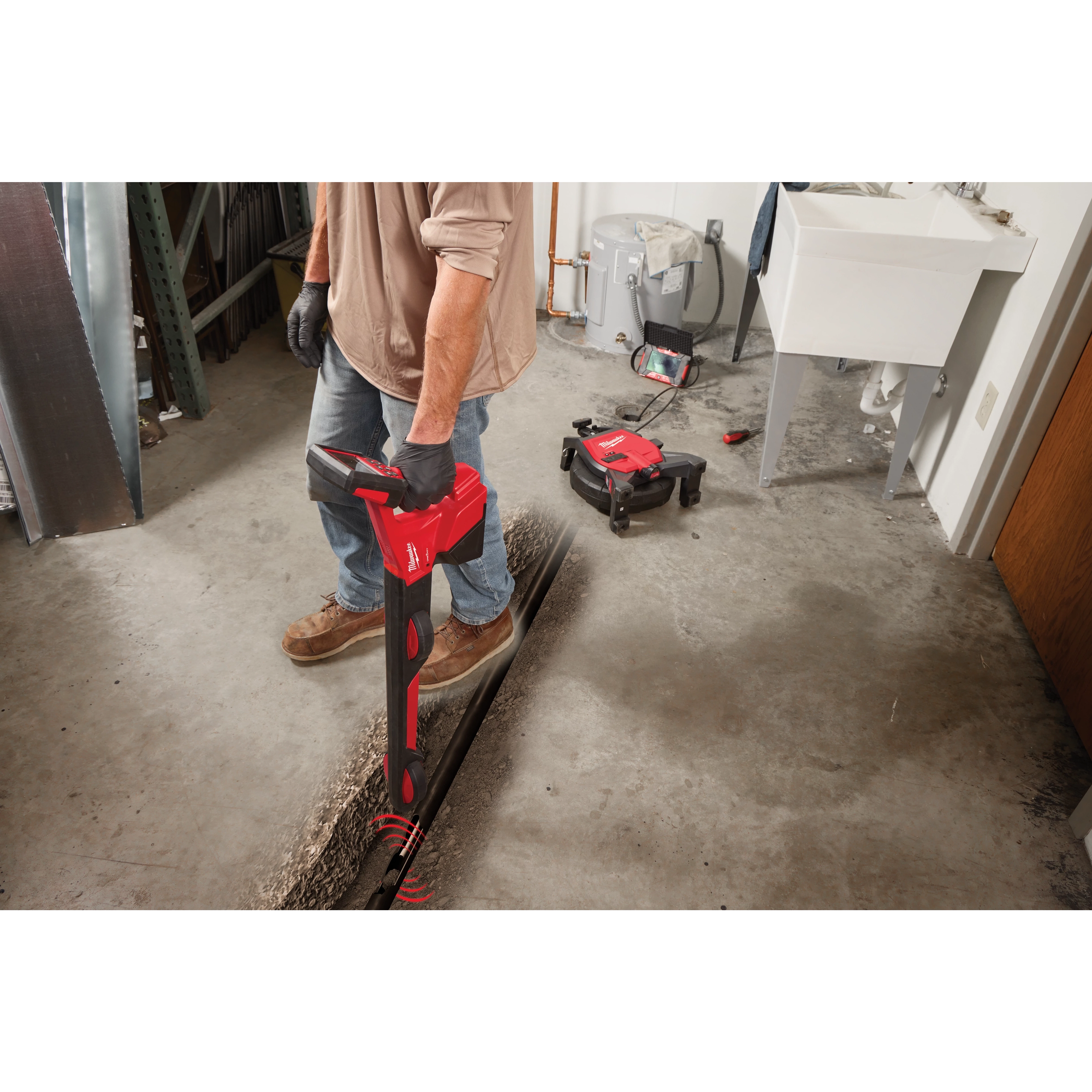 A person uses a red pipe locator to detect underground pipes in a concrete floor. Nearby are tools including a remote controlled device, various repair tools, and a white sink. The person wears gloves, blue jeans, and work boots.