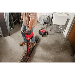 A person uses a red pipe locator to detect underground pipes in a concrete floor. Nearby are tools including a remote controlled device, various repair tools, and a white sink. The person wears gloves, blue jeans, and work boots.
