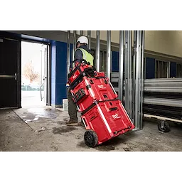 Worker with hard hat and safety vest moving PACKOUT XL Tool Box stack on wheels through a construction site doorway.
