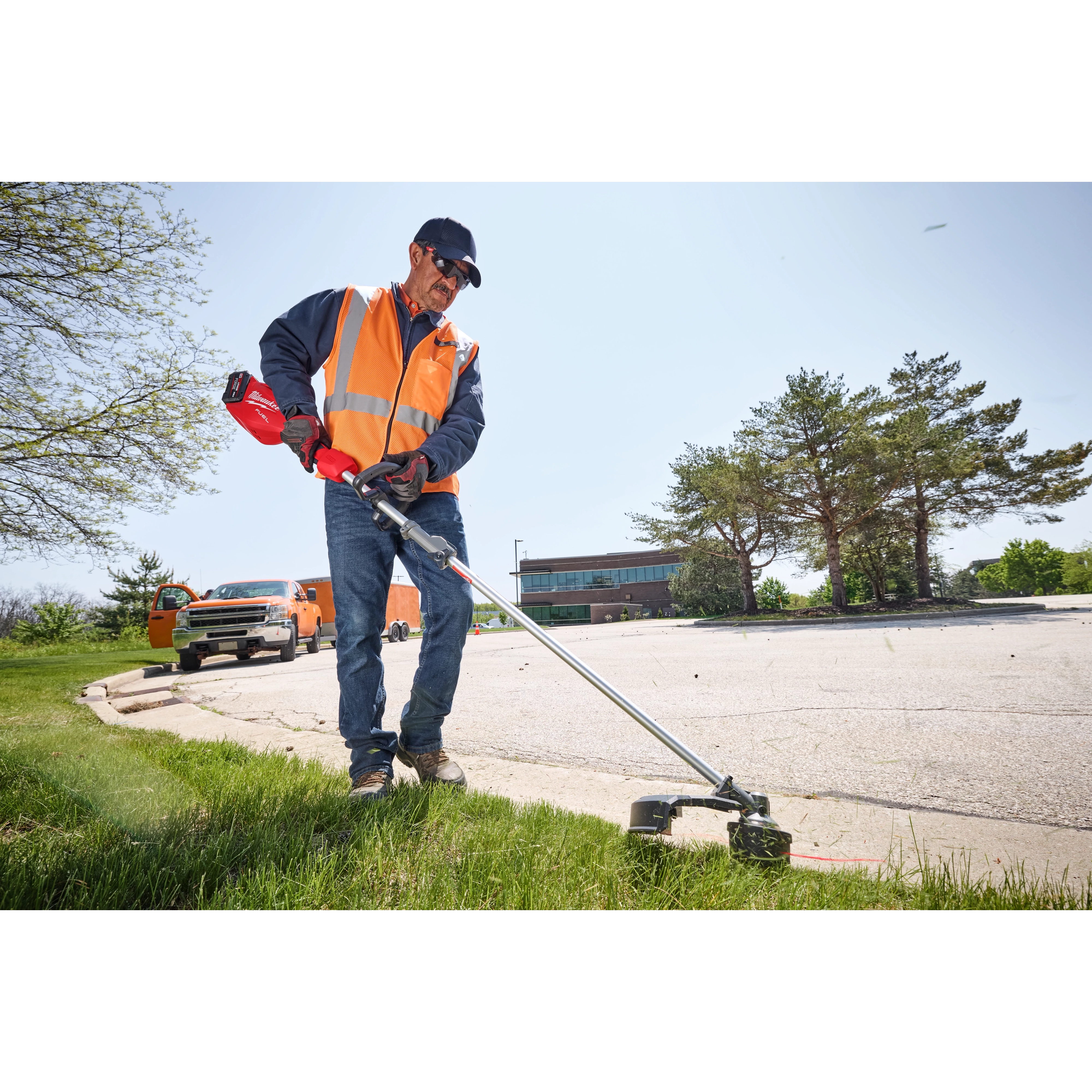 A person wearing an orange vest and using an M18 FUEL™ Power Head w/ QUIK-LOK™ trimmer adjusts the grass edge on a lawn near a parking lot with trees and vehicles in the background. The person stands on a grassy area edging along the pavement.