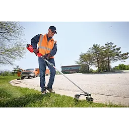 A person wearing an orange vest and using an M18 FUEL™ Power Head w/ QUIK-LOK™ trimmer adjusts the grass edge on a lawn near a parking lot with trees and vehicles in the background. The person stands on a grassy area edging along the pavement.