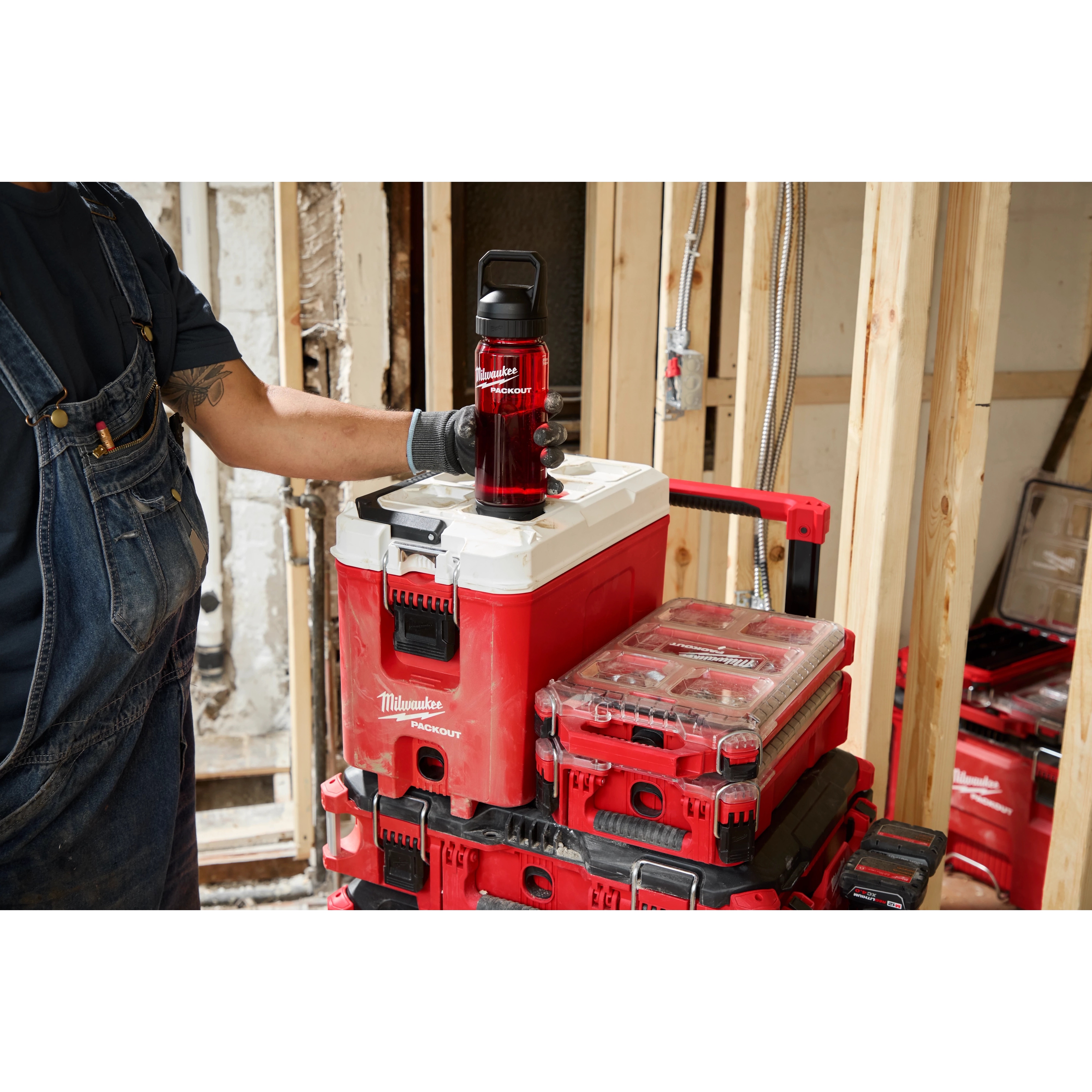 A person wearing overalls and gloves places a red PACKOUT™ 25oz Bottle with Chug Lid on top of a red and white PACKOUT™ storage box. More PACKOUT™ storage boxes are stacked on a trolley in a construction environment.