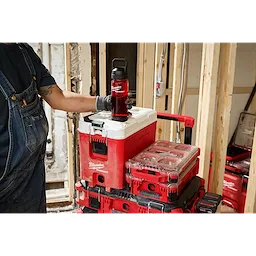 A person wearing overalls and gloves places a red PACKOUT™ 25oz Bottle with Chug Lid on top of a red and white PACKOUT™ storage box. More PACKOUT™ storage boxes are stacked on a trolley in a construction environment.