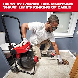 A person operates the Milwaukee drain snake tool in a bathroom, feeding the cable into a floor drain. The drain snake is mounted on a wheeled cart with cable storage and has a handle for ease of use. The text above reads, "Up to 3x longer life: maintains shape, limits kinking of cable."