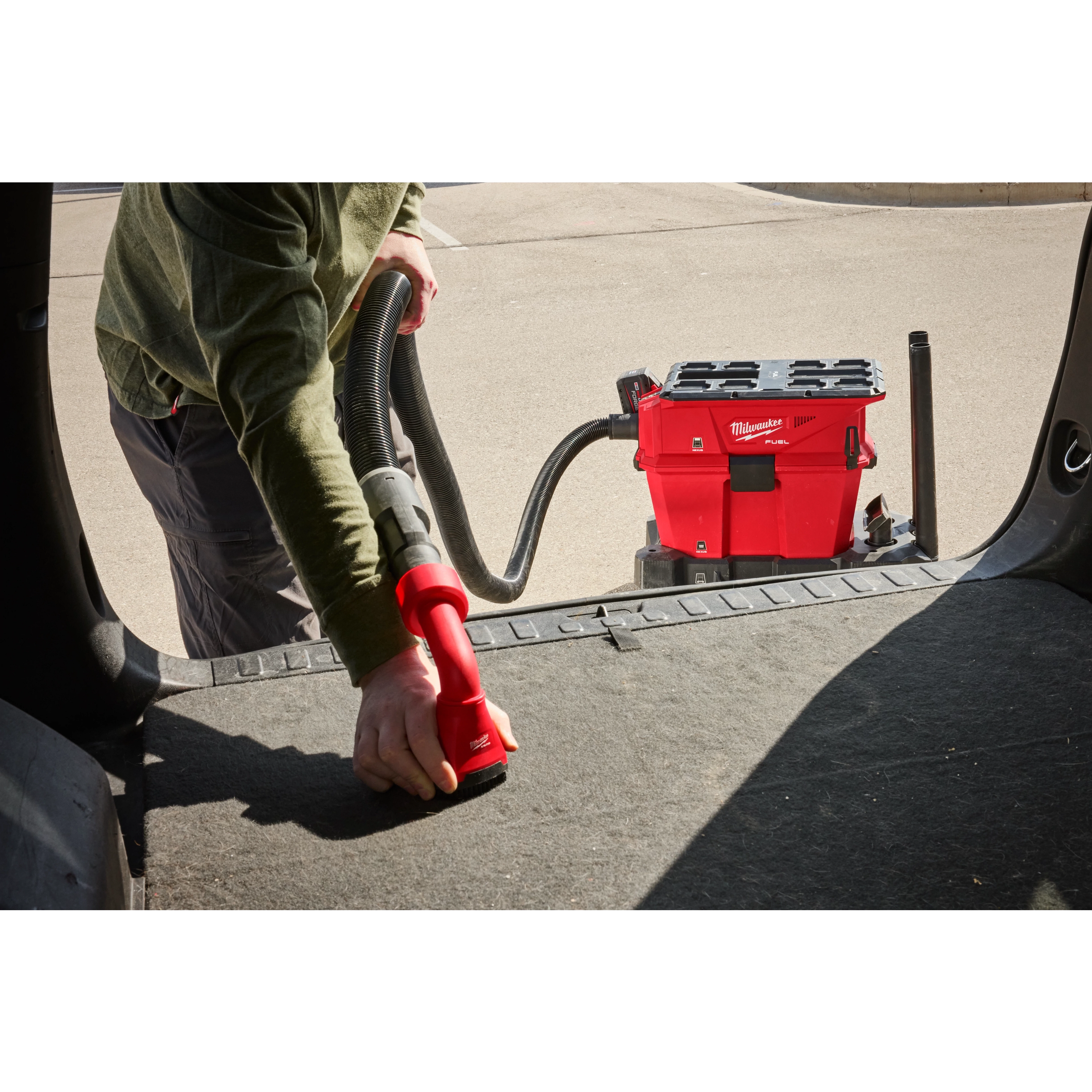 A person uses the M18 FUEL™ NEXUS™ 6 Gallon Wet/Dry Vacuum w/ PACKOUT™ Compatibility to clean the interior of a vehicle. The red vacuum is positioned on the ground, and the person is handling the vacuum hose inside a vehicle.