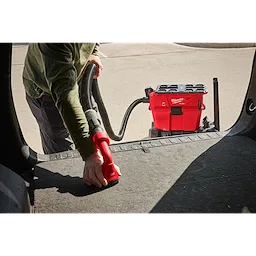 A person uses the M18 FUEL™ NEXUS™ 6 Gallon Wet/Dry Vacuum w/ PACKOUT™ Compatibility to clean the interior of a vehicle. The red vacuum is positioned on the ground, and the person is handling the vacuum hose inside a vehicle.