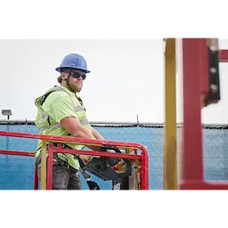 A construction worker in a safety vest, hard hat, and harness stands on a cherry picker. The worker is operating the machine at a worksite. They wear Full Frame Safety Glasses with Removable Side Shields and Tinted Anti-Scratch Lenses. The background features a blue safety net and fence.