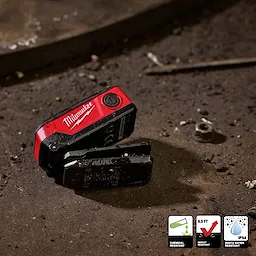 Milwaukee pocket flood light lying on a dusty metal surface with small debris nearby.