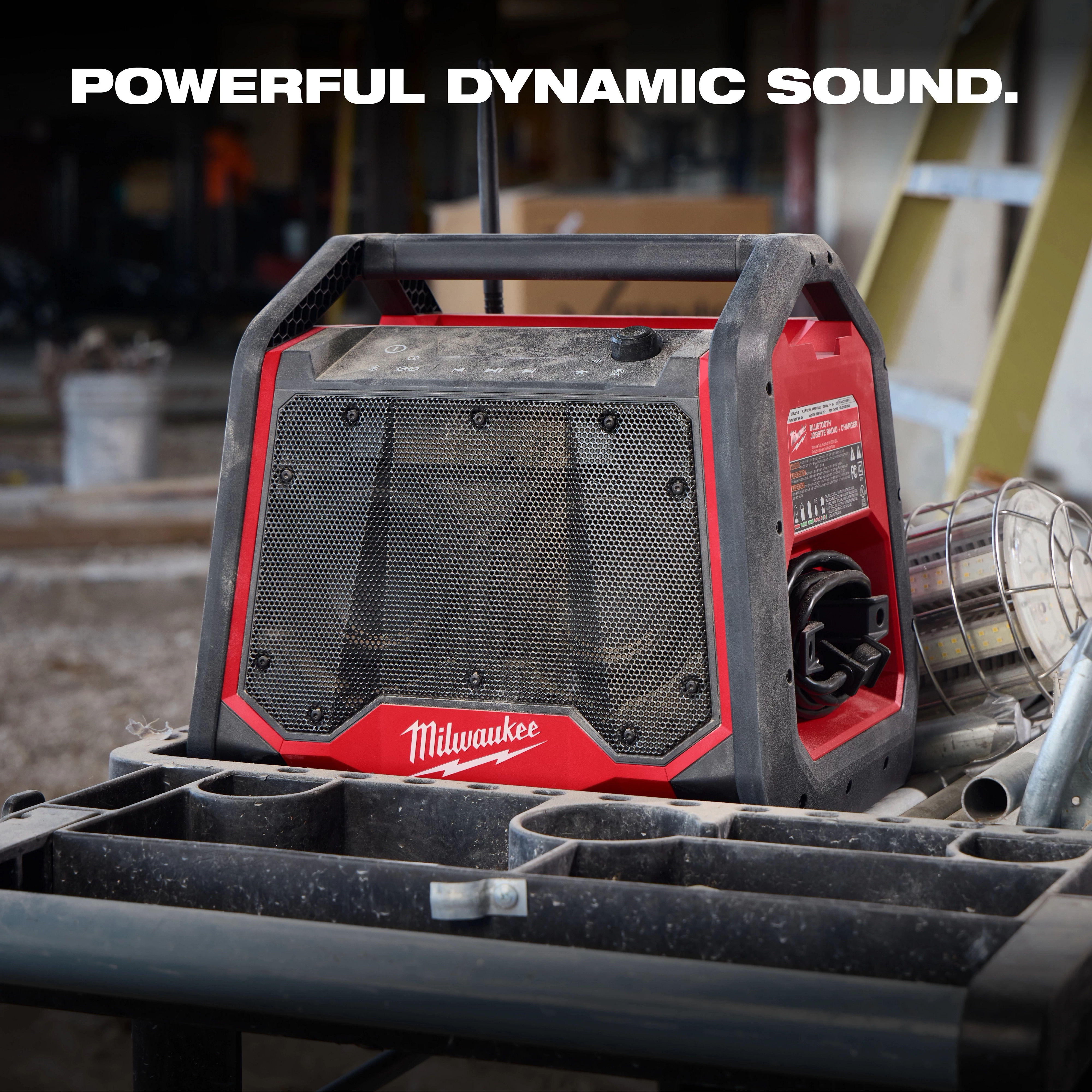 The M18 BLUETOOTH JOBSITE RADIO + CHARGER is red and black with a rugged design. This radio is placed on a construction site workbench. Bold text at the top reads “POWERFUL DYNAMIC SOUND.”