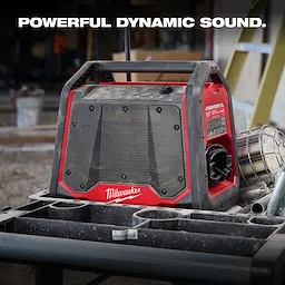 The M18 BLUETOOTH JOBSITE RADIO + CHARGER is red and black with a rugged design. This radio is placed on a construction site workbench. Bold text at the top reads “POWERFUL DYNAMIC SOUND.”