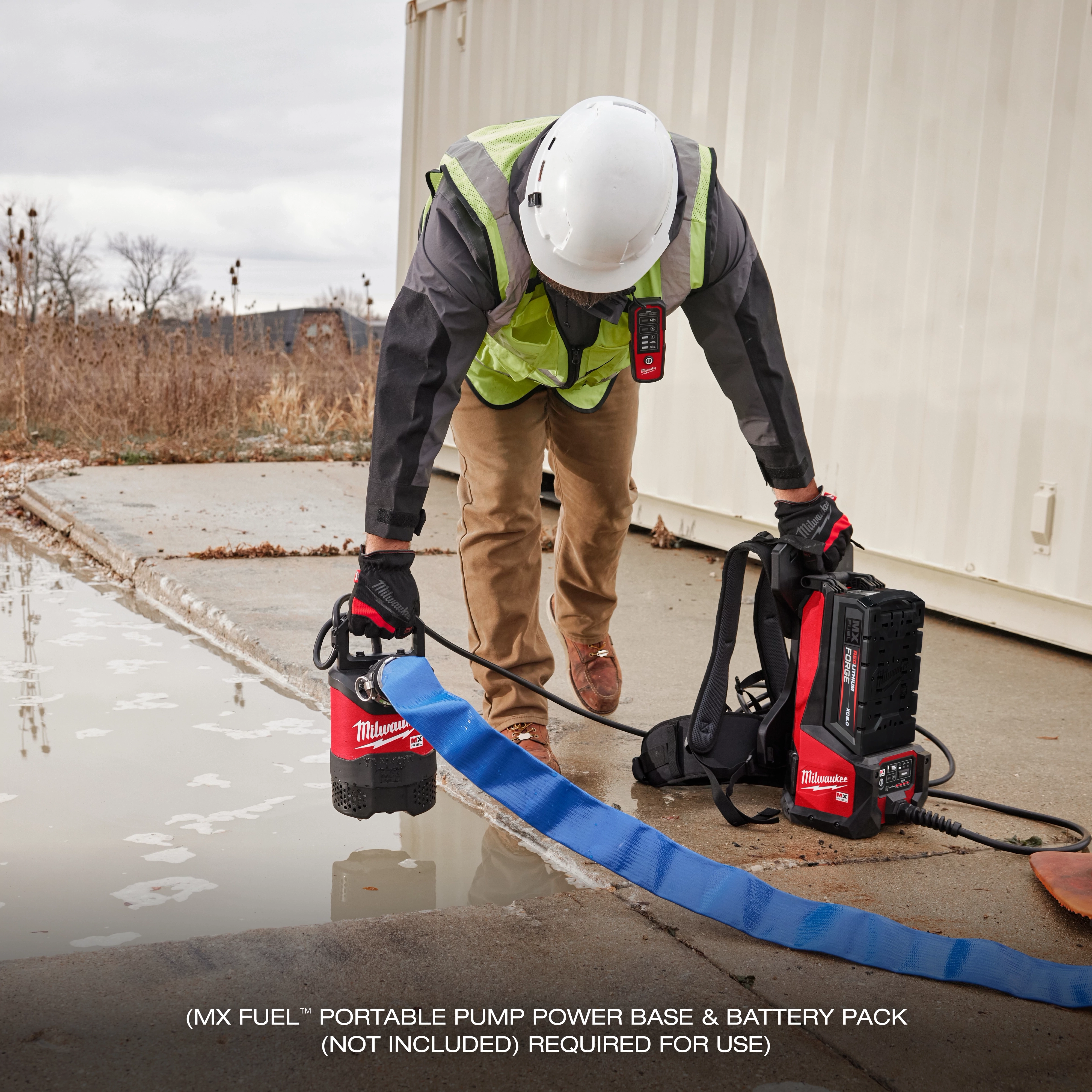 A person wearing a hard hat and safety vest operates an MX FUEL™ 1HP 2" Submersible Pump next to a shallow puddle. The pump is connected to a blue hose and a red and black power base. The text indicates a portable pump power base and battery pack are required but not included.