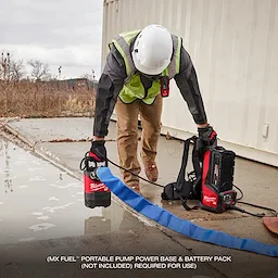 A person wearing a hard hat and safety vest operates an MX FUEL™ 1HP 2" Submersible Pump next to a shallow puddle. The pump is connected to a blue hose and a red and black power base. The text indicates a portable pump power base and battery pack are required but not included.