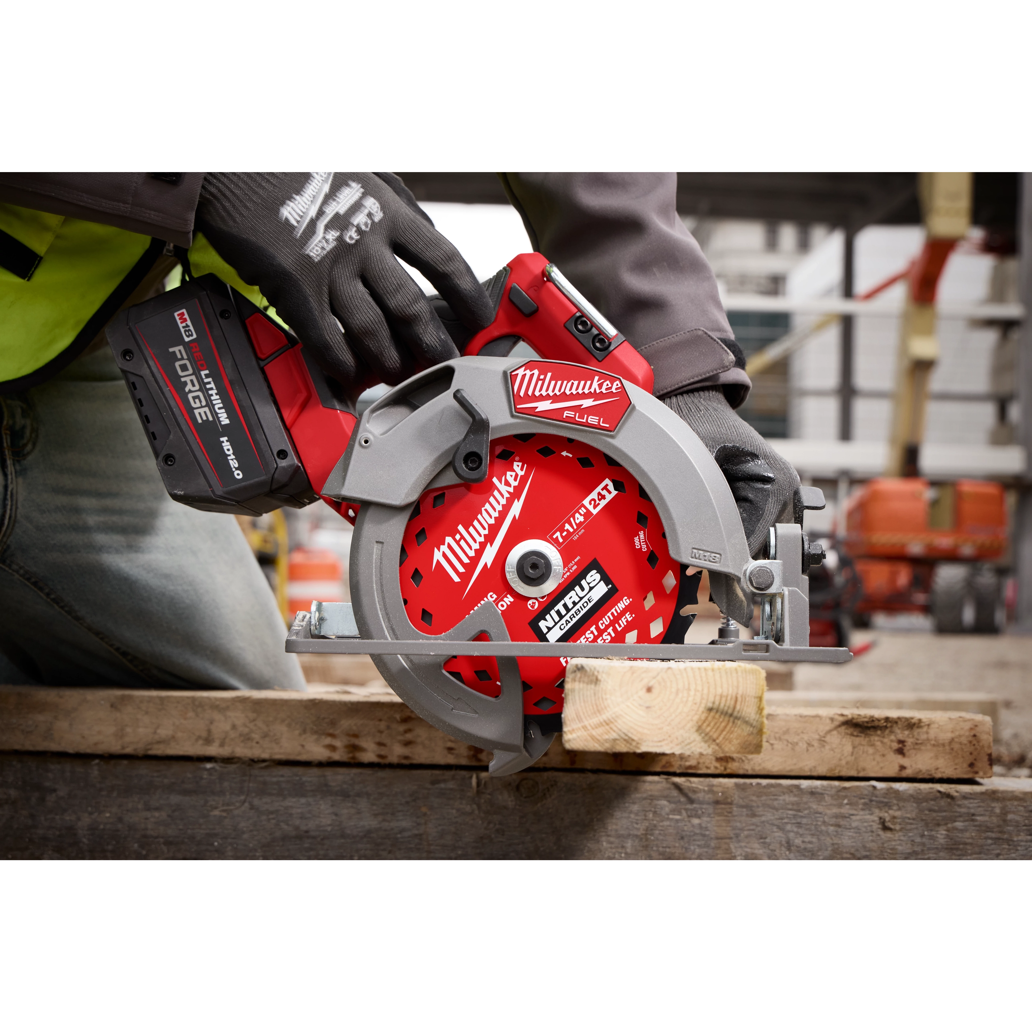 A construction worker wearing safety gloves uses a red Milwaukee Fuel circular saw to cut a wooden plank. The saw is angled downwards on the wood, and the scene appears to be at an active construction site with blurred background elements like scaffolding and materials.