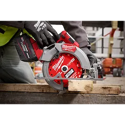 A construction worker wearing safety gloves uses a red Milwaukee Fuel circular saw to cut a wooden plank. The saw is angled downwards on the wood, and the scene appears to be at an active construction site with blurred background elements like scaffolding and materials.