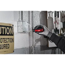A gloved hand uses a #2 Phillips 4" Cushion Grip Screwdriver (USA) to adjust a screw on an electrical box mounted on a gray wall. A yellow "Caution" sign is visible on the left side of the image.