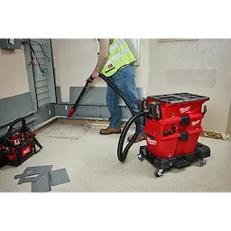 A worker uses the M18™ FUEL™ NEXUS™ Dual Battery Wet/Dry Vacuum Motor Head with PACKOUT™ Compatibility & VACLINK™ to clean up a work area. The red vacuum with black hoses and attachments is positioned on a wheeled base for mobility. Nearby, a tool bag and debris are visible on the floor.