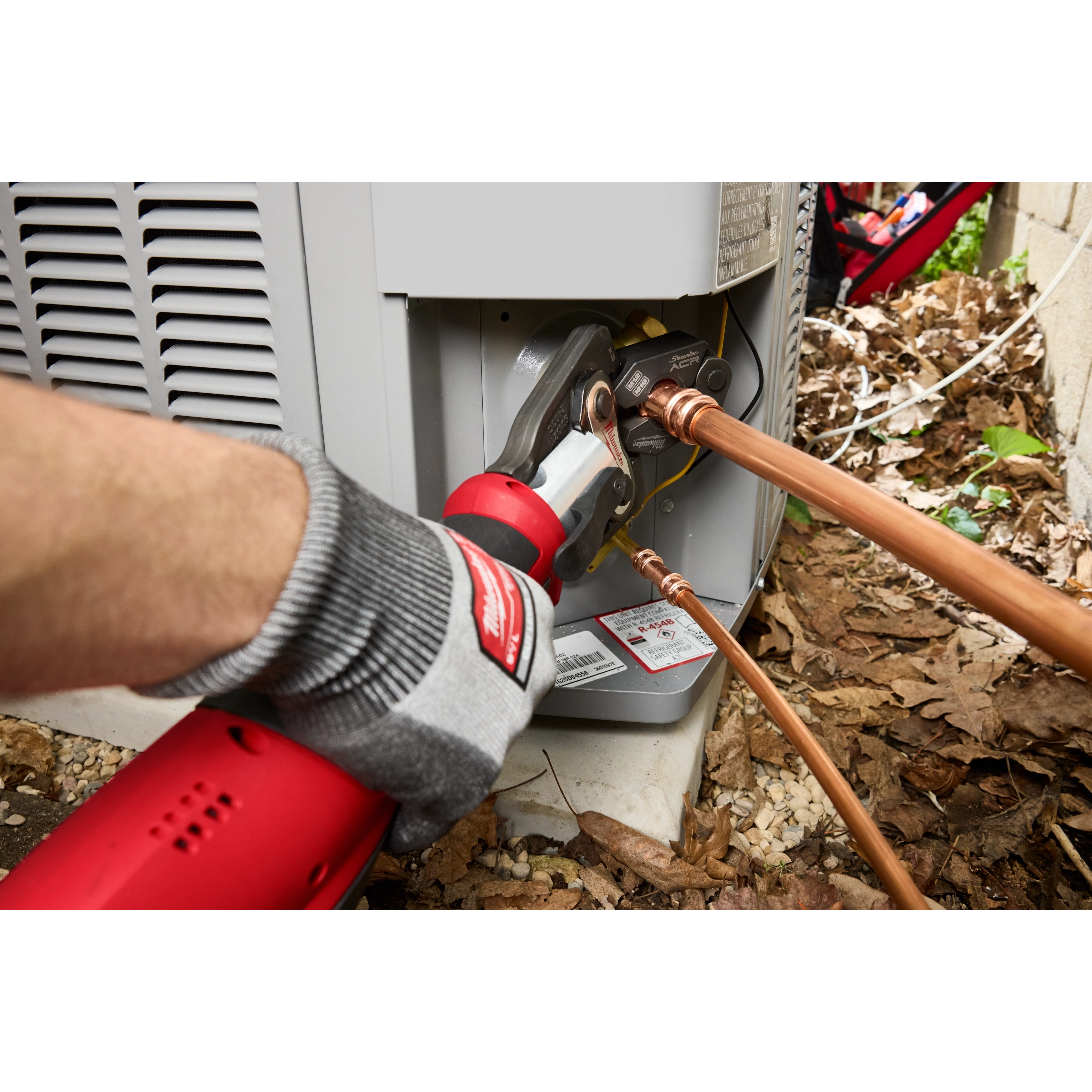 The Milwaukee ProPress tool connects copper pipes to an outdoor HVAC unit. An angled view shows a gloved hand operating the red and silver device. The unit exterior, leaves, and debris are visible in the background, providing context for the installation environment.