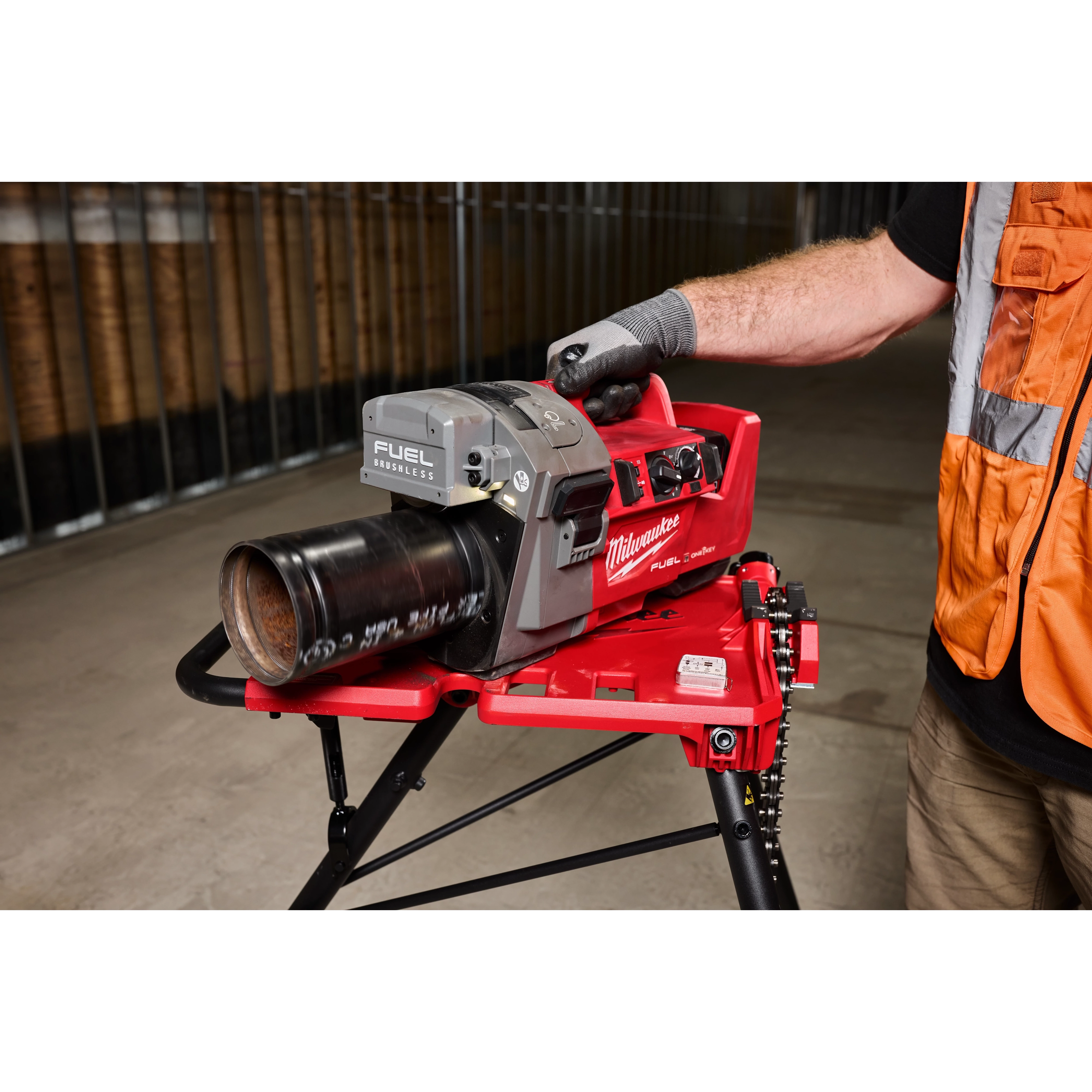 A person operates an M18 FUEL™ RINGER™ Roll Groover for 2” – 6” Sch 10/40 on a red stand, indoors. The tool is marked with "Milwaukee" branding and is used for grooving metal pipes. The person is wearing gloves and a safety vest.