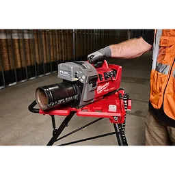 A person operates an M18 FUEL™ RINGER™ Roll Groover for 2” – 6” Sch 10/40 on a red stand, indoors. The tool is marked with "Milwaukee" branding and is used for grooving metal pipes. The person is wearing gloves and a safety vest.