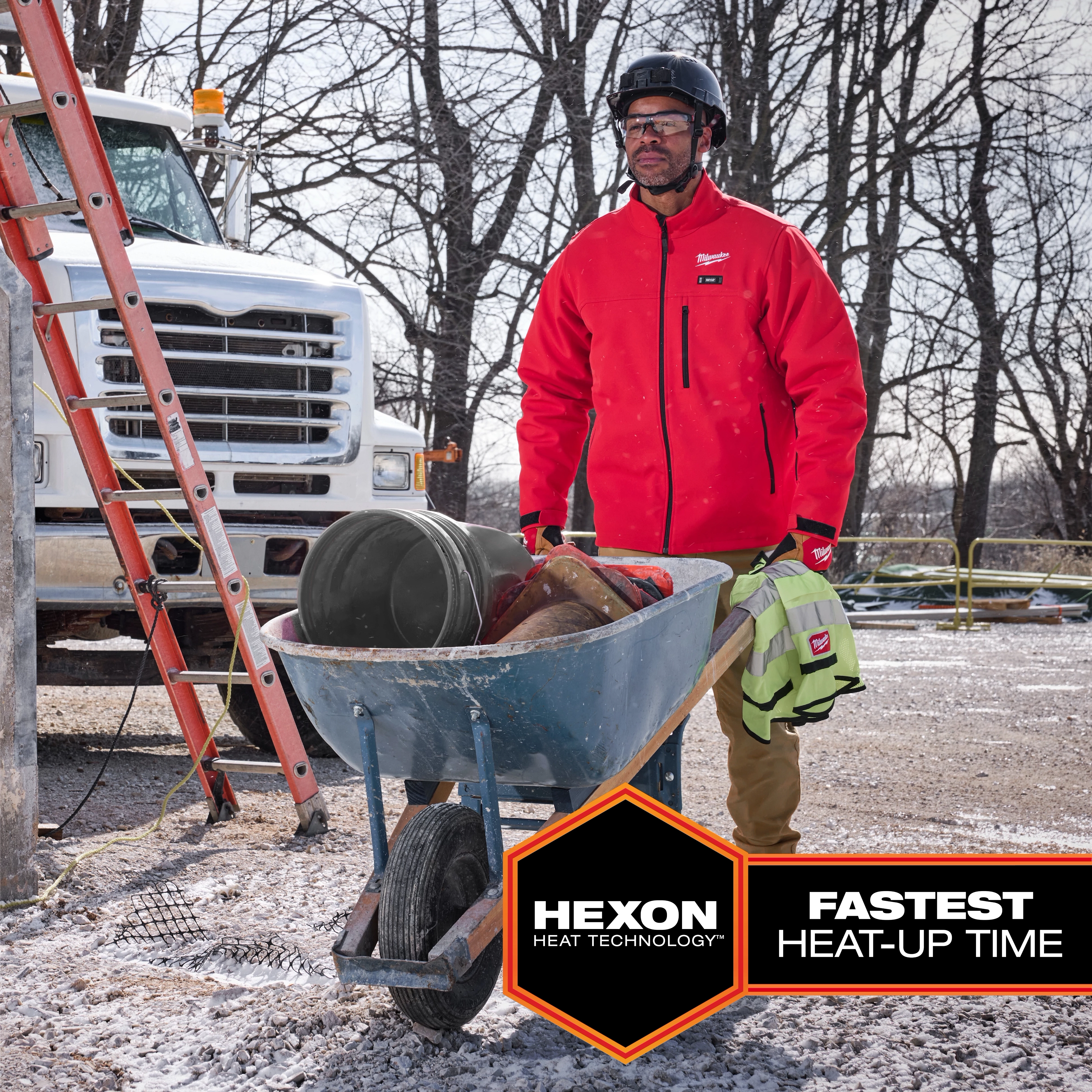 Worker wearing a red M12 Heated TOUGHSHELL Jacket while carrying a wheelbarrow filled with tools on a construction site.