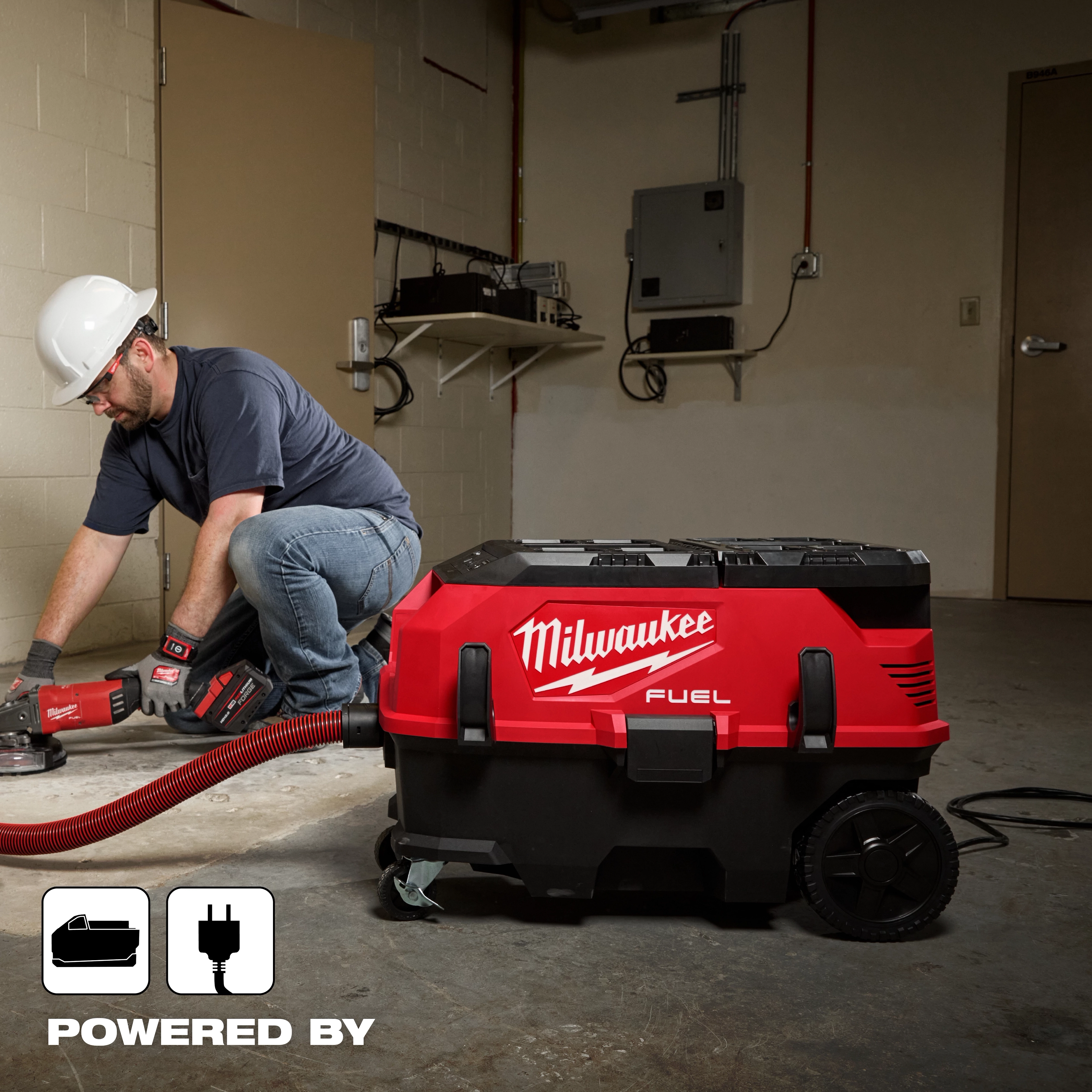 A worker uses the Milwaukee M18 FUEL™ 9 Gallon Dual Battery Dust Extractor w/ PACKOUT™ Compatibility and VACLINK™ in a workshop. The dust extractor is red and black, with large wheels and a flexible hose connected to a power tool.