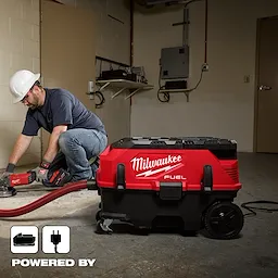 A worker uses the Milwaukee M18 FUEL™ 9 Gallon Dual Battery Dust Extractor w/ PACKOUT™ Compatibility and VACLINK™ in a workshop. The dust extractor is red and black, with large wheels and a flexible hose connected to a power tool.