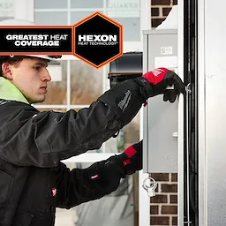 A person wearing black Milwaukee-branded Heated Work Glove Liners is seen working on an electrical box. The image highlights the gloves' "Greatest Heat Coverage" and "Hexon Heat Technology."