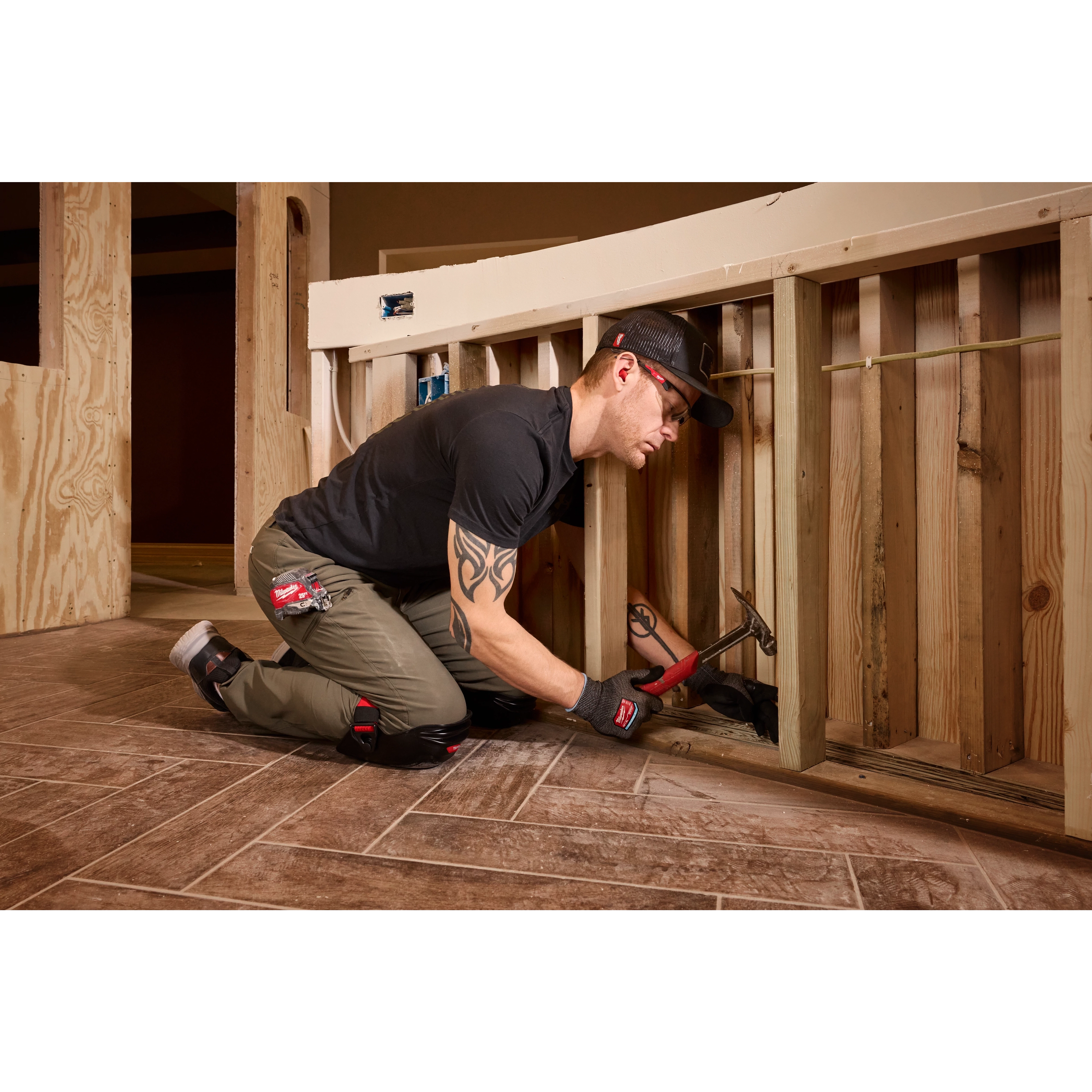 A person wearing a Snapback Bump Cap is kneeling and working on a wooden construction frame. They are using a hammer and gripping a piece of wood in a partially built structure.
