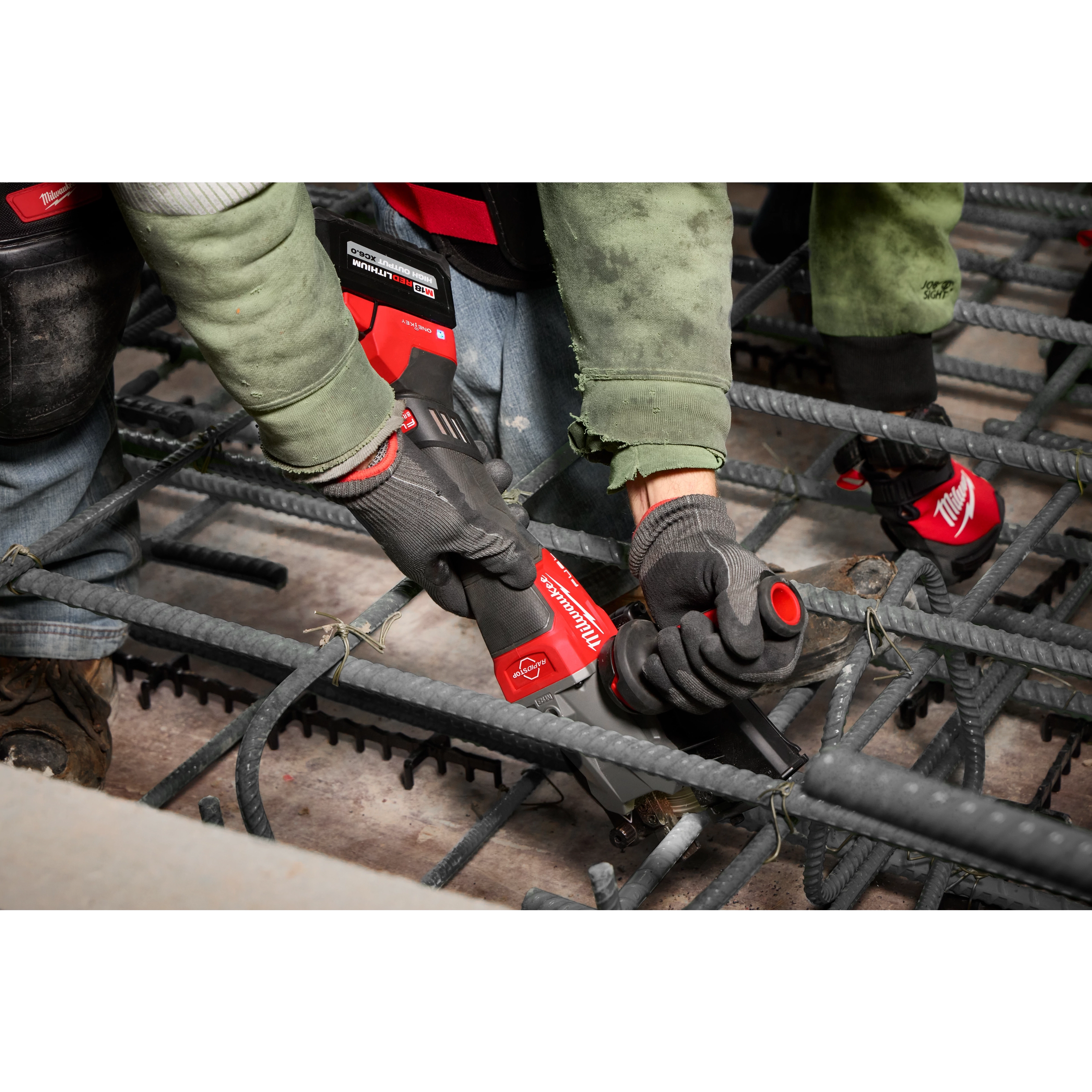 Two construction workers are using the M18 FUEL™ #10 (1-1/4") Rebar Cutter to cut steel rebar. The rebar cutter is red and black, and the workers are wearing gloves and protective clothing while working on a concrete rebar structure.