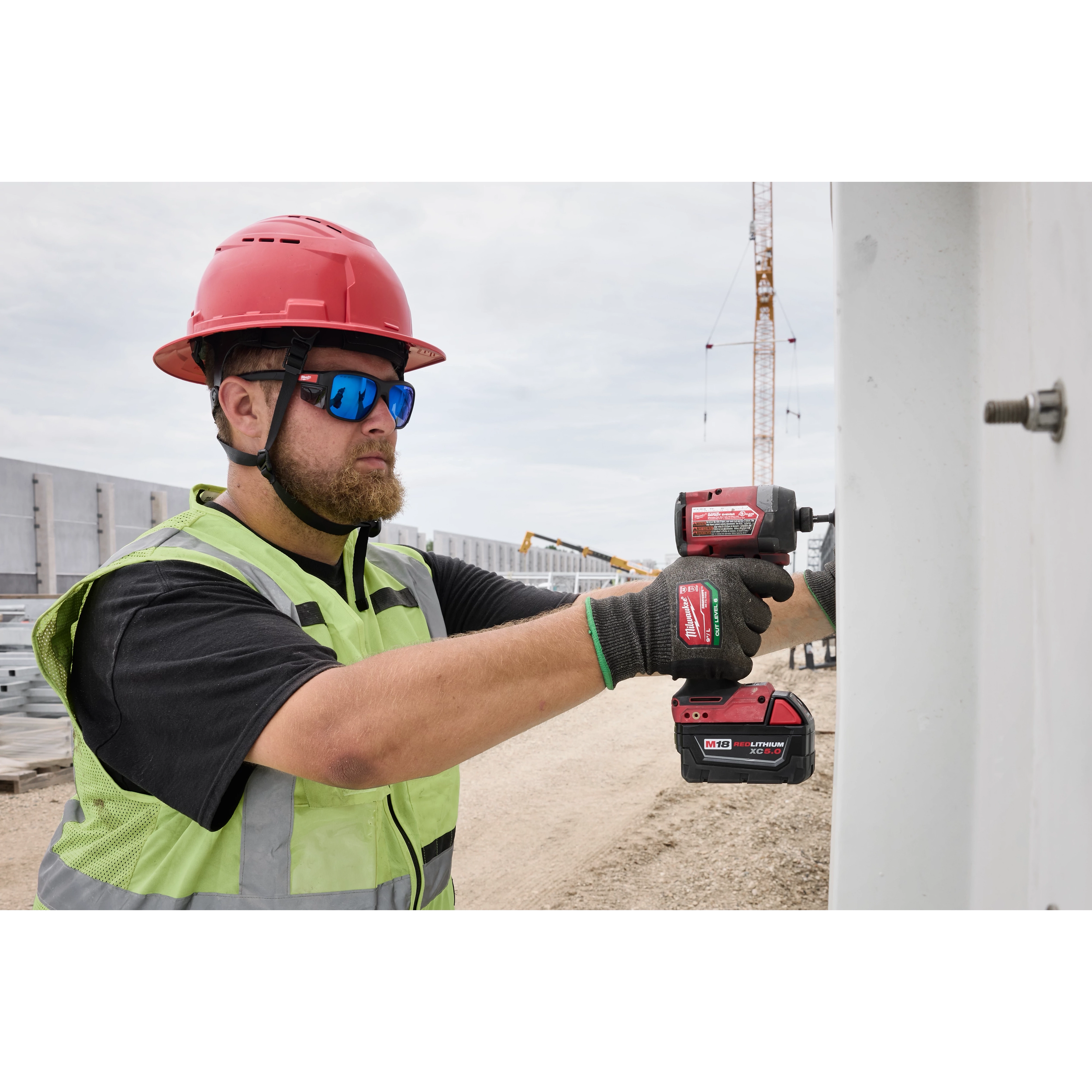 A construction worker in a red hard hat and fluorescent safety vest uses a battery-operated impact driver to secure a bolt. The worker is wearing Polarized Full Frame Safety Glasses with Removable Side Shields – Blue Mirrored Dual Coat Lenses, providing robust eye protection in an outdoor work environment.