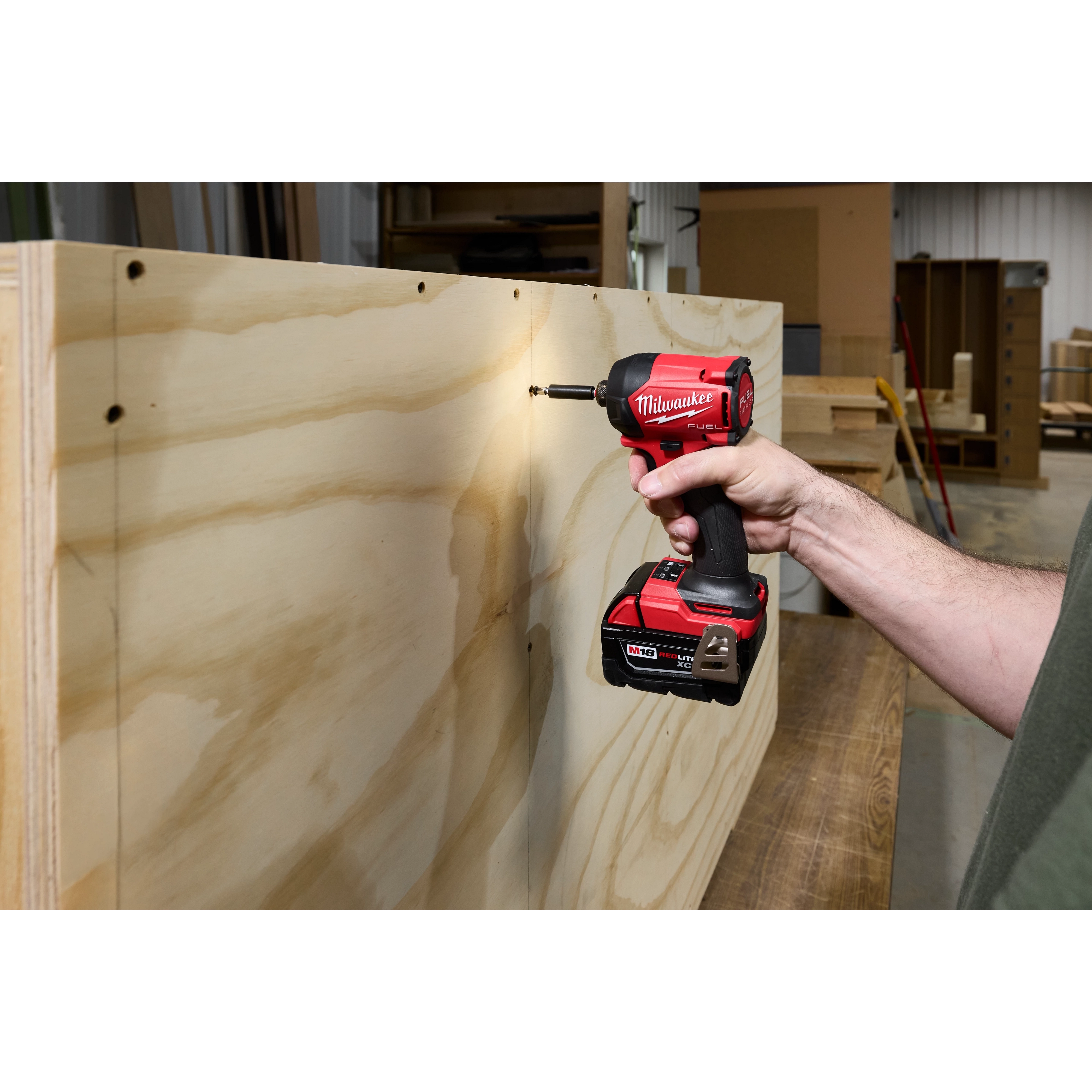 A person is using the M18 FUEL™ SURGE™ 1/4" Hex Hydraulic Driver, a red and black power tool, to drive a screw into a wooden board in a workshop setting.