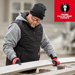 A person is wearing a Milwaukee M12 Heated AXIS Vest in black, with a grey hoodie underneath and red work gloves. The product features secure full-zip front pockets. They appear to be working with tools and materials at an outdoor construction site.